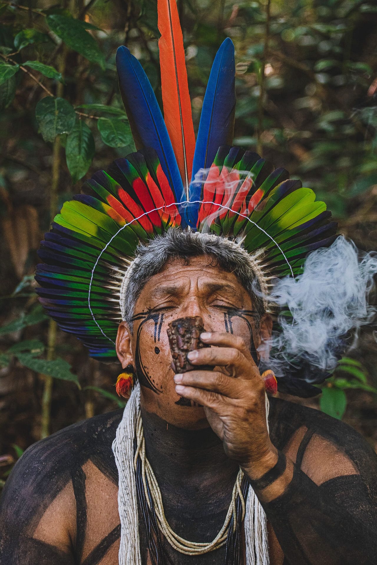 Portrait of an indigenous man with traditional attire and smoke in a Brazilian forest.